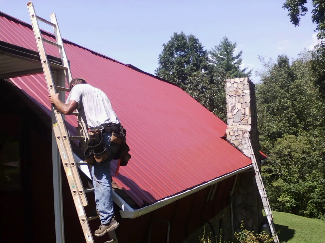 Roofer on a ladder beside a red metal roof and stone chimney — Soffit & Fascia Repair by Roofing Brothers in Margaretville